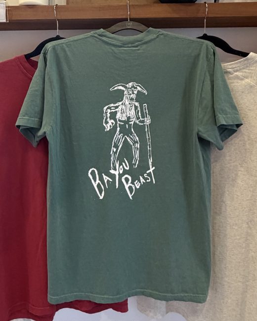 The Bayou Beast t-shirt in sage green on the merch rack, with the hand drawn beast printed on the back