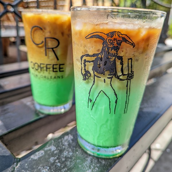 Two Bayou Beast iced lattes on the patio rail, coffee brown layered over bright pandan green, with a hand drawn horned beast on the front glass and the CR Coffee logo on the glass behind