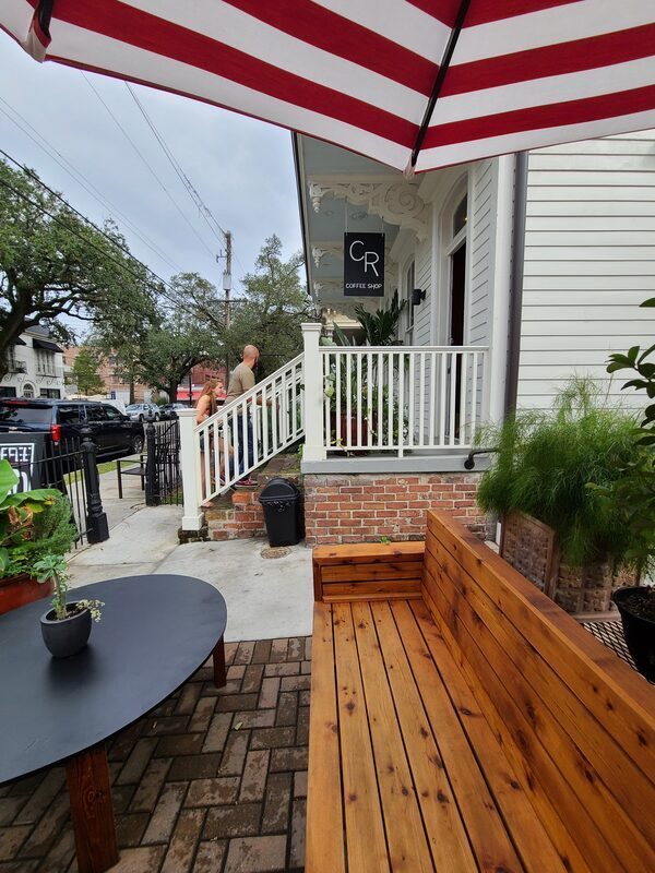 The side patio: a cedar bench and cafe table under a red and white striped umbrella, beside the white steps and CR Coffee Shop sign at the Magazine Street entrance