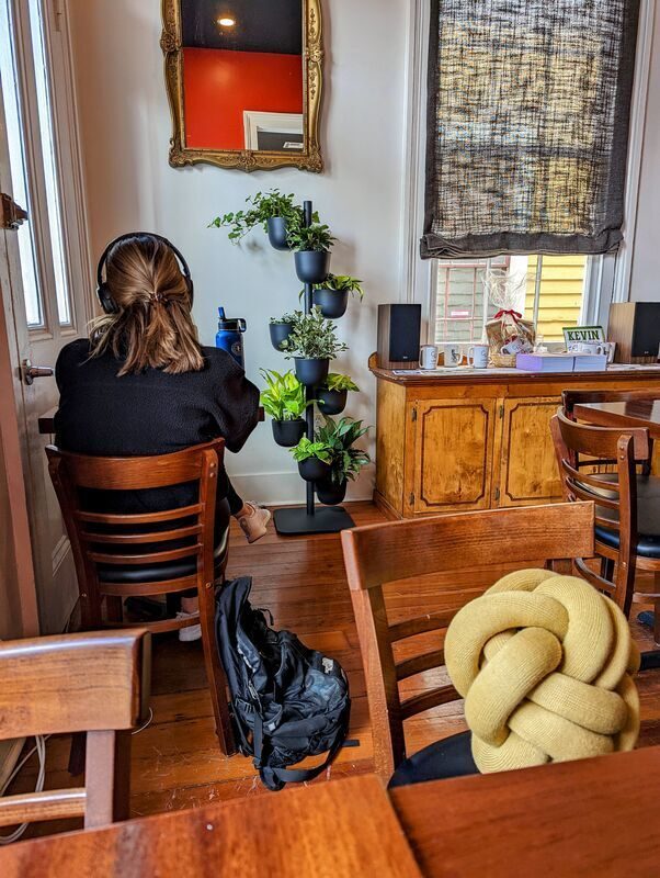 A student in headphones working at a table inside the Magazine Street shop, backpack on the wood floor beside her, with a wall of potted plants and a gold framed mirror behind