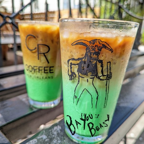 Two Bayou Beast iced lattes on a rail, the front glass showing a hand drawn horned beast above the words Bayou Beast, with bright green pandan settled at the bottom