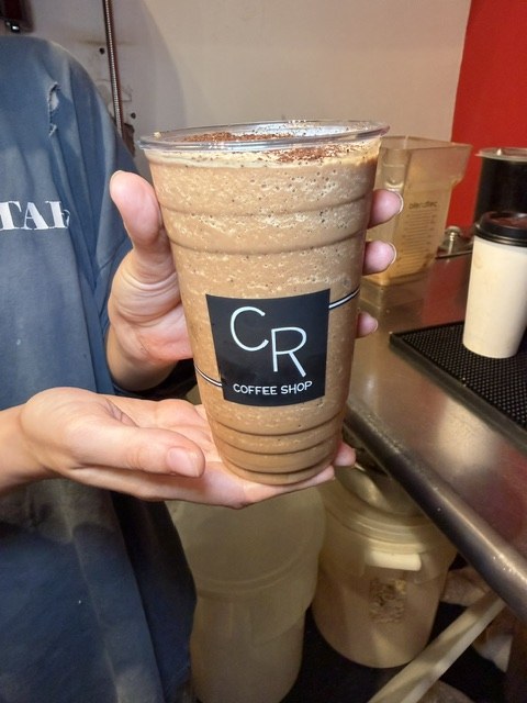 The same frozen drink held up behind the bar, dark espresso grounds covering the whole surface and flecks of ground coffee suspended through the blended coffee below