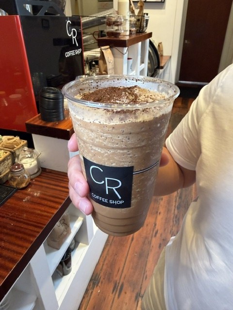 A tall clear CR Coffee Shop cup held in one hand at the Magazine Street counter, filled with thick light brown frozen coffee and dusted heavily with dark espresso grounds on top
