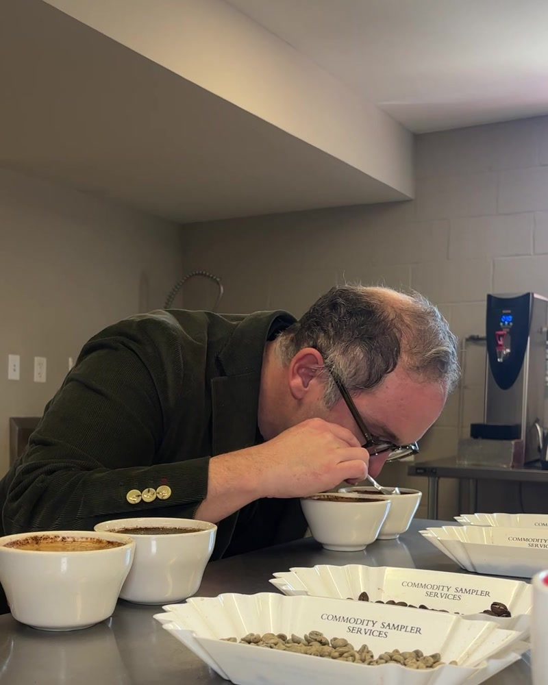 Kevin leans over the cupping table, nose close to a ceramic bowl, breaking the crust and evaluating the aroma of a fresh sample