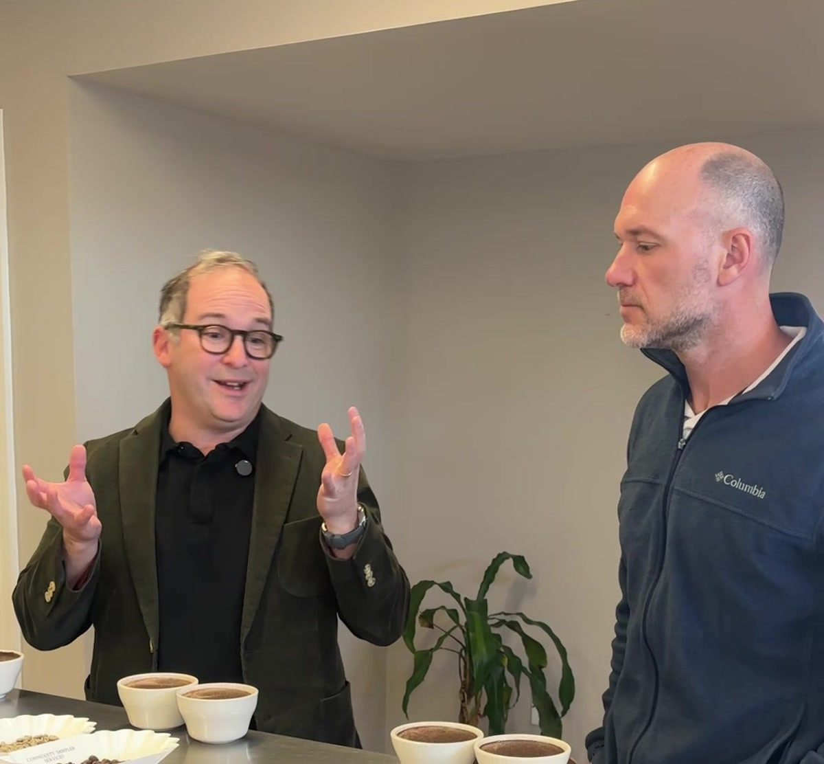 Kevin gestures while discussing coffee samples with an importer at the cupping table, ceramic cups and bean trays lined up in front of them