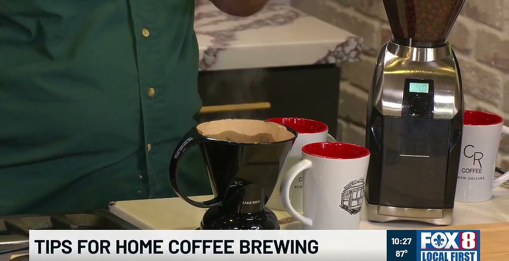 A black Clever Coffee Dripper steaming on the Fox 8 Kitchen counter next to two CR Coffee mugs with red interiors and a Baratza burr grinder full of beans