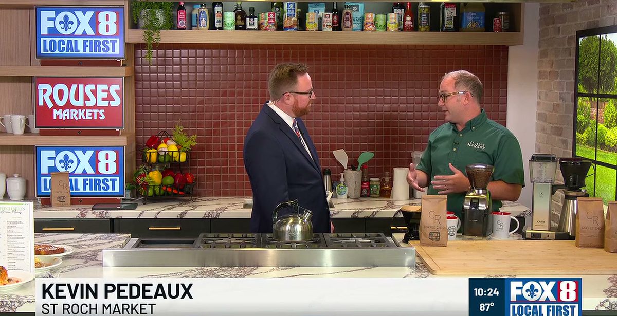 Kevin Pedeaux in a green St. Roch Market polo talking with anchor Rob Krieger at the Fox 8 Kitchen counter, with CR Coffee bags, mugs, a burr grinder, and a drip machine laid out and an on-screen banner reading Kevin Pedeaux, St. Roch Market