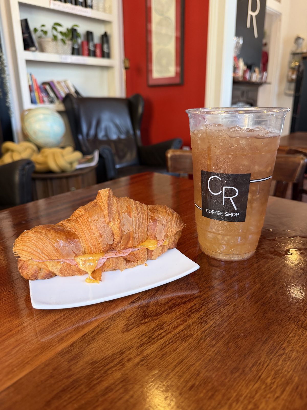 A ham and cheese croissant on a white plate with melted cheese running out the side and pooling on the plate, next to a tall iced tea in a clear CR Coffee Shop cup, on a polished wood table with the red walls and armchairs of the shop behind it