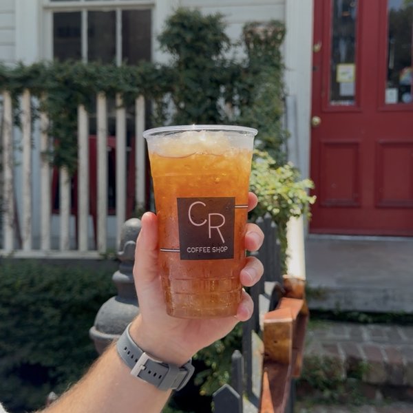 A large iced tea in a clear CR Coffee Shop cup held up in front of the white porch and red front door of the Magazine Street shop