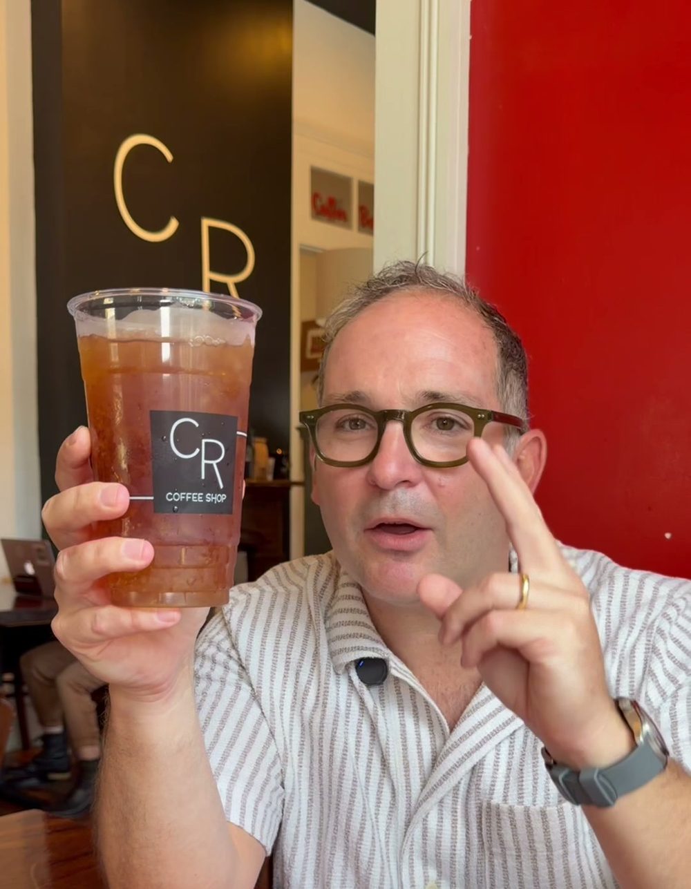 Kevin Pedeaux holding up a large iced tea in a CR Coffee Shop cup inside the Magazine Street shop, gesturing with one hand while talking