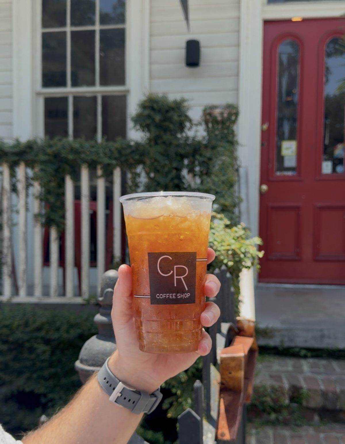 A large Lagniappe Iced Tea in a clear CR Coffee Shop cup held up in front of the white porch and red front door of the Magazine Street shop
