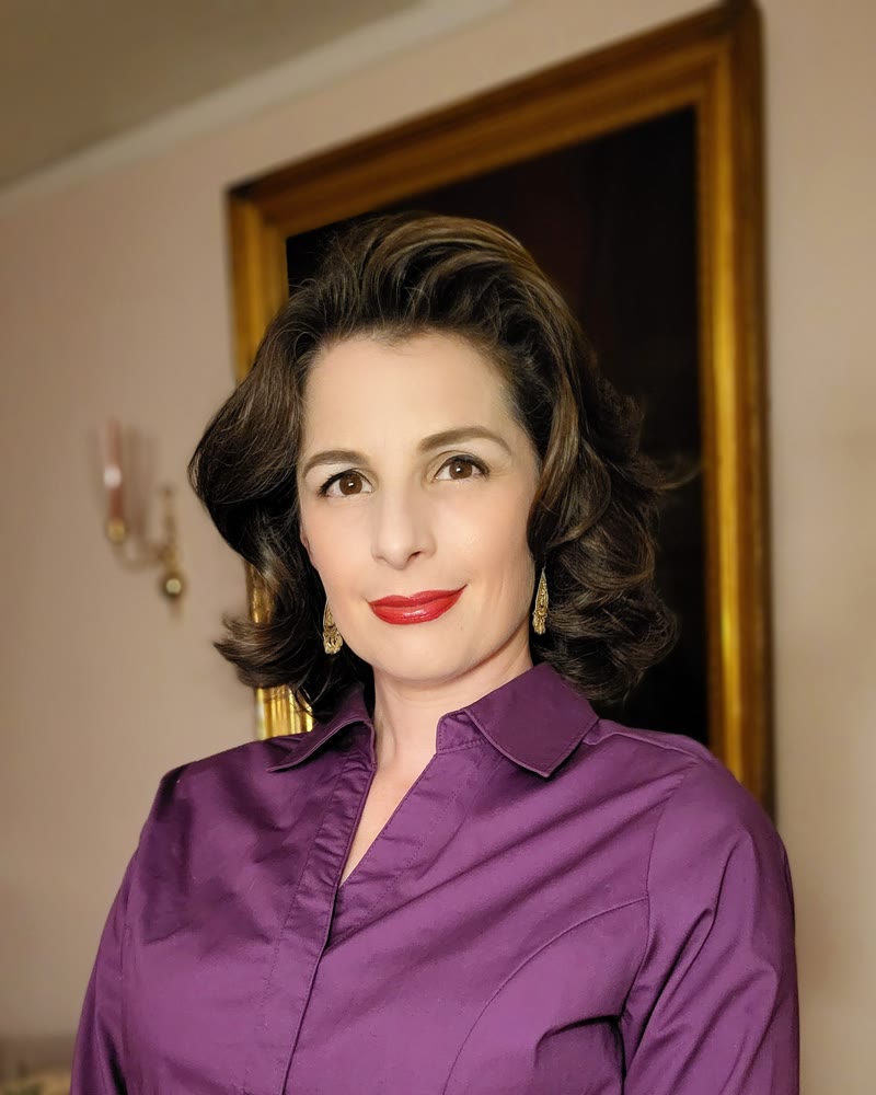 Portrait of author Marielle Songy in a purple collared shirt, smiling, in front of a gilt-framed mirror