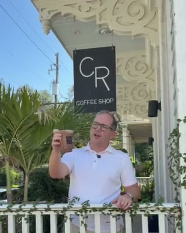 Kevin Pedeaux holding up a Protein Cold Brew on the front porch of CR Coffee Shop on Magazine Street, under the black CR Coffee Shop sign