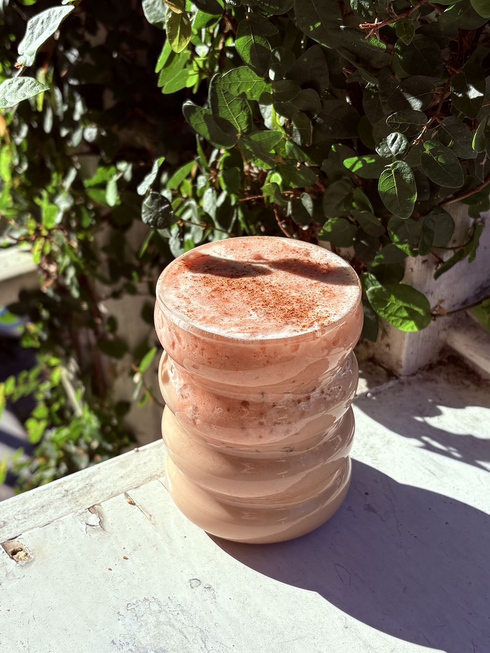 Iced Strawberry Chai sitting on a white wood railing on the patio at CR Coffee Shop, surrounded by green leaves in spring sunlight
